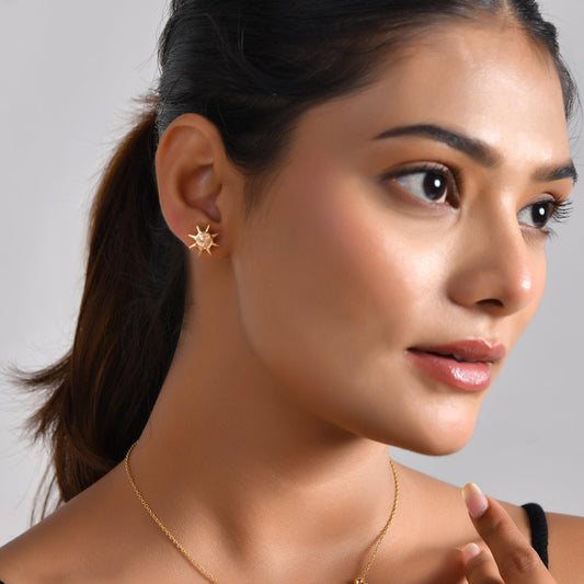 A woman with dark hair tied back wears a delicate gold necklace with small colorful charms and a matching gold star-shaped stud earring, posed against a neutral gray background.