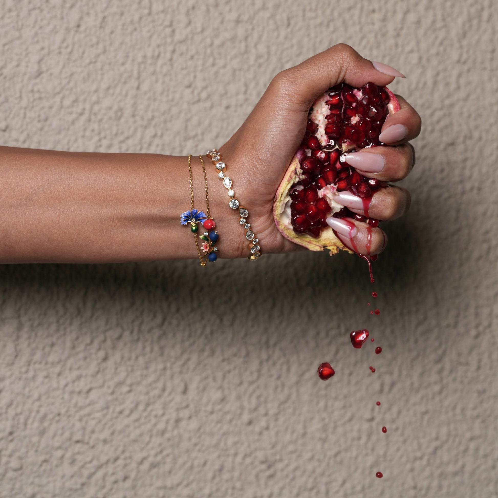 Hand squeezing a pomegranate against a beige wall wearing gold bracelet