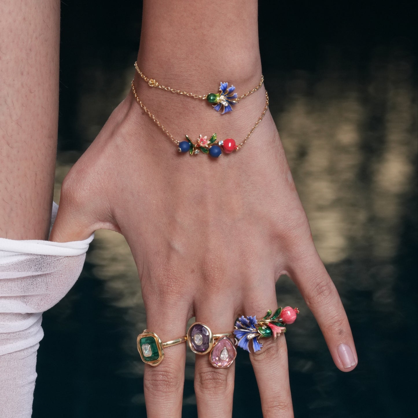 Hand wearing a gold bracelet and rings with colorful gemstones against a blurred natural background