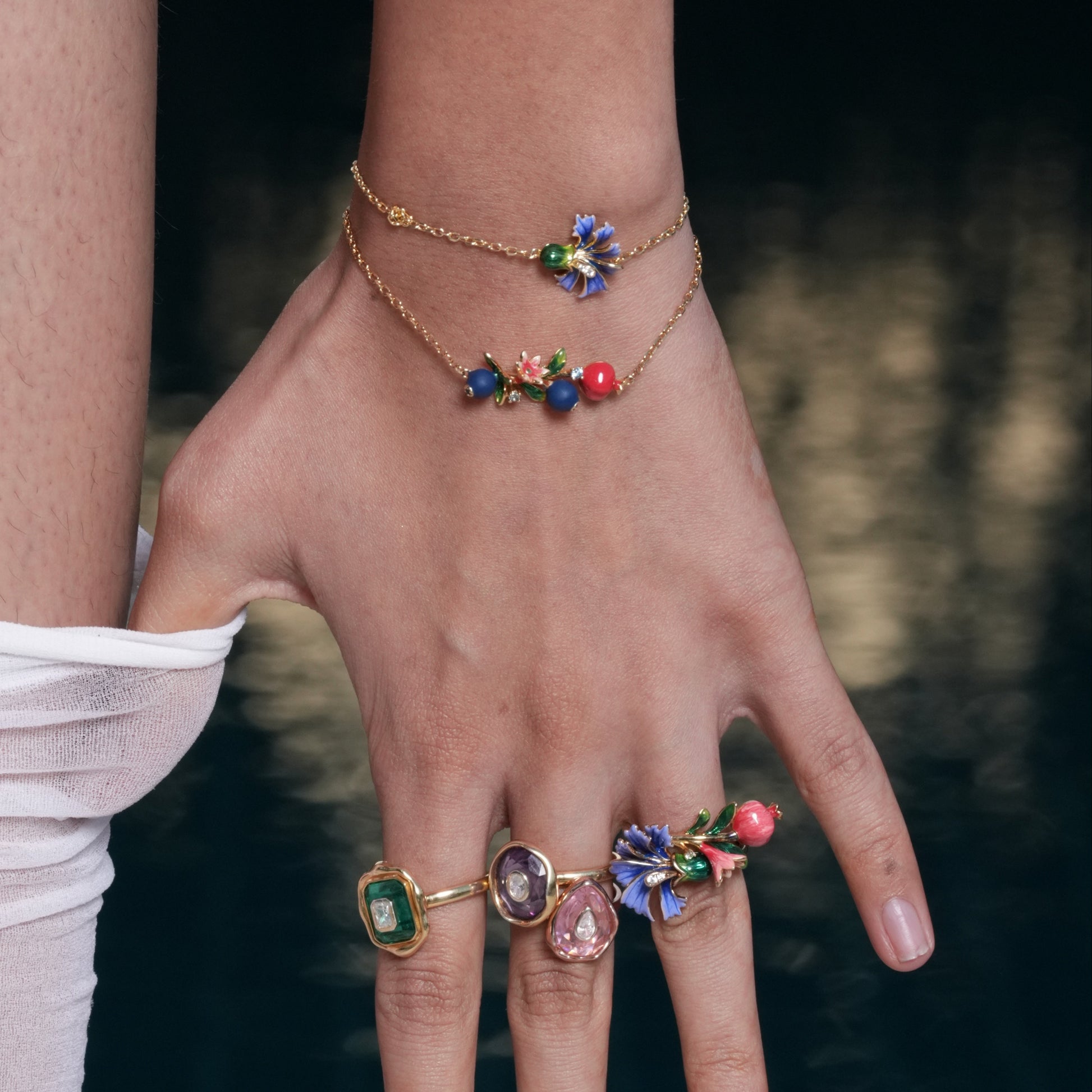 Hand wearing a gold bracelet and rings with colorful gemstones against a blurred natural background